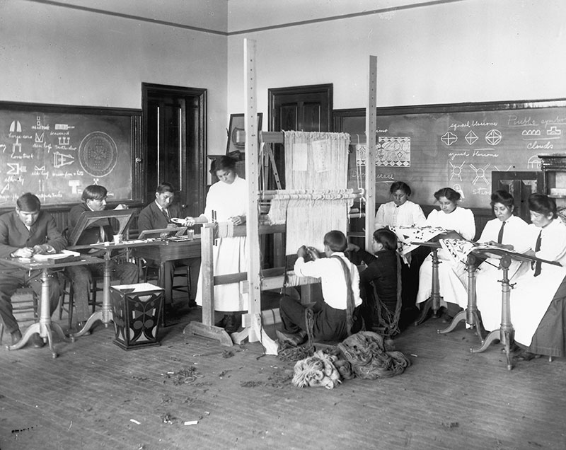 Early 1900s black and white photo of Native youth in an art class weaving and working with fibers