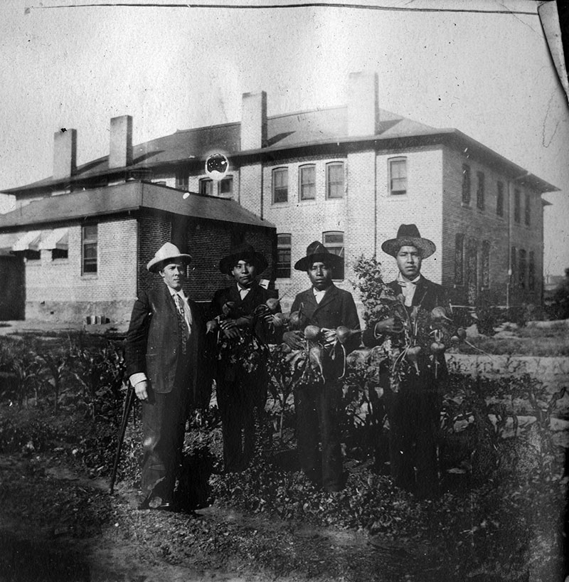 1904 black and white photo of 3 Native youth holding armfuls of turnips next to an older white man in front of a large 2-story brick building
