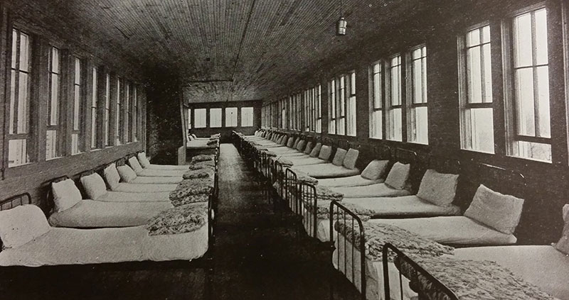 1918 black and white photo of a long narrow wood floor and ceilng room with many windows and single beds lined along the walls.