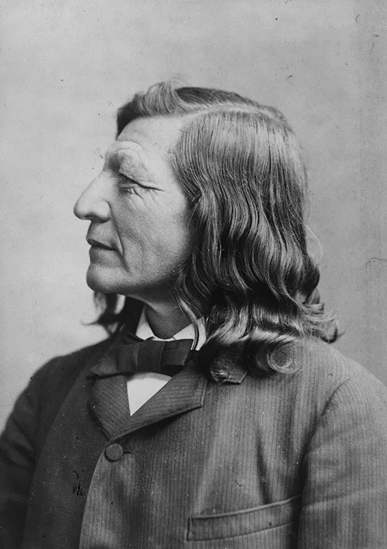 Black-and-white portrait of a man with shoulder-length hair in a suit, facing left. He has a prominent nose and a serious expression.