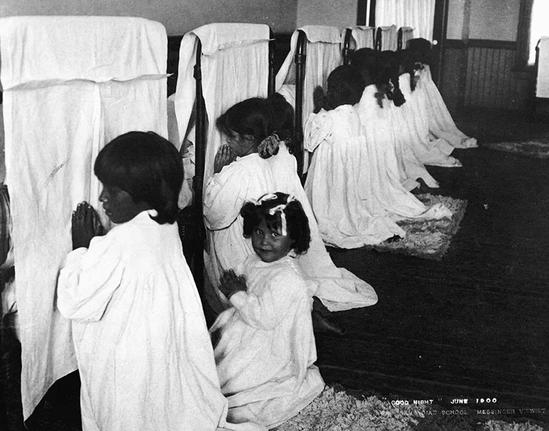 A group of young children in nightgowns kneel in prayer beside their beds in a dormitory, with dividers between each bed. The photo is dated June 1908.