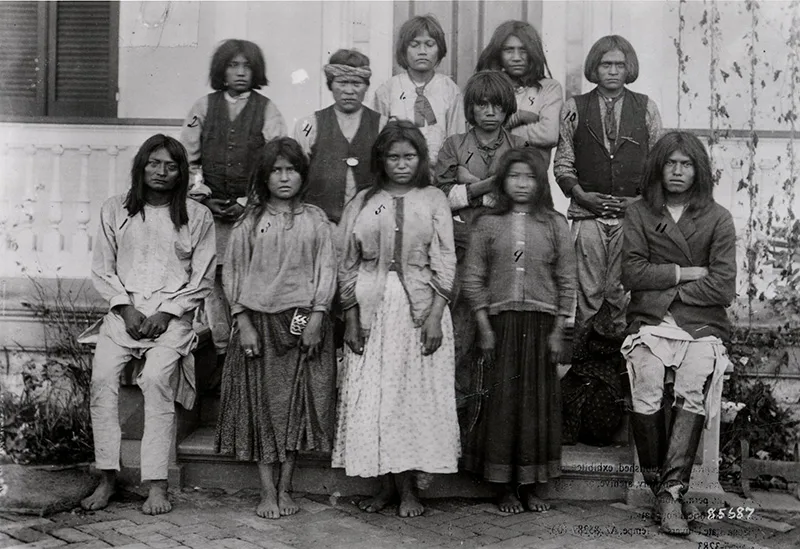A historic black and white photo of a group of 11 Apache individuals, some seated and others standing, in front of a building. Each person has a number labeled on them.