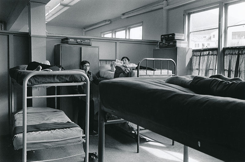 1970 black and white photo of large dorm hall with multiple metal framed bunk beds with 2 young men. One is lounging on an upper bunk reading and the other stands next to the bed.