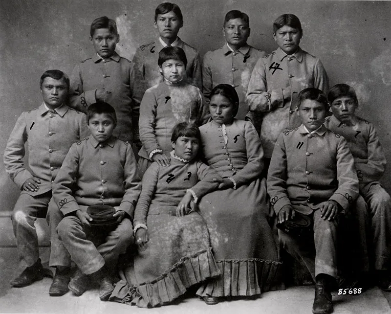 Black-and-white photograph of a group of Native American students in uniforms from the Carlisle Indian Industrial School, posing in two rows, with seven boys standing and four girls seated in the front.
