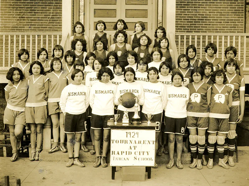 1929 black and white photo of large group of young Native women from 4 different basketball teams. They pose for the camera in different uniforms with trophies on a table and a banner saying "1929 Tournament at Rapid City Indian School" in the foreground.