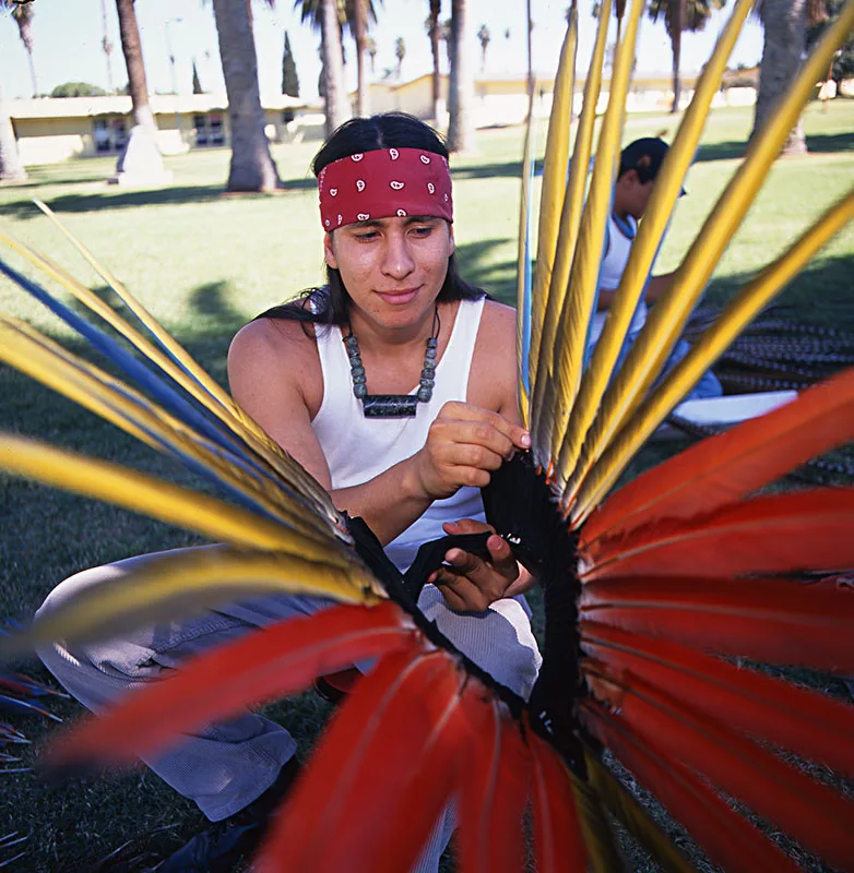 1999 candid color photo of a young Native man wearing a red bandana around his forehead and carefully aligning long feathers on a head dress