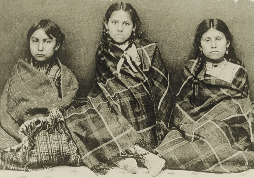 Three young girls sit side by side, wrapped in patterned blankets, looking directly at the camera in a historical black-and-white photograph.