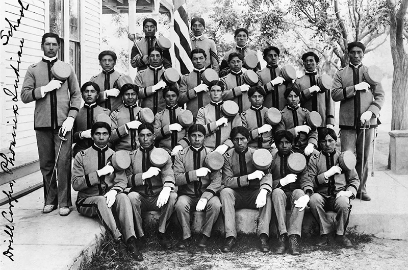 Early 20th C. black and white photo of young Native men in military style uniforms with caps in their hands and many over their hearts posing for a group portrait.