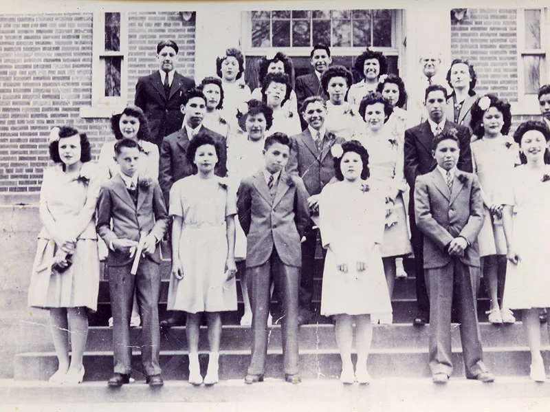 1940 black and white photo of young Native men and women dresses and suits posing on steps leading up to a brick building.