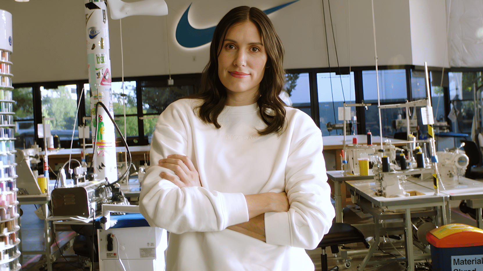 A person with long hair stands confidently with crossed arms in a modern workspace filled with sewing machines and equipment. The Nike logo is visible in the background.