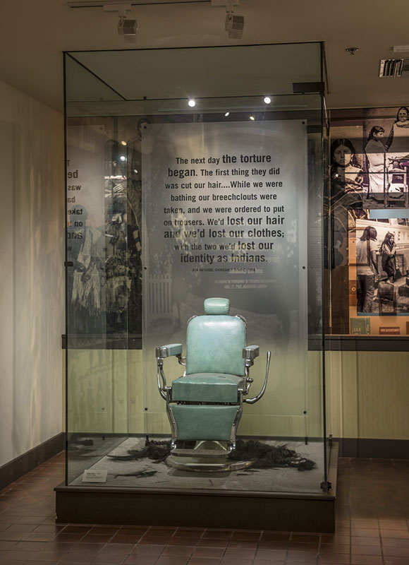 A vintage barber chair is displayed in a glass case. Text on the back wall describes the historical context of forced haircuts and loss of identity.