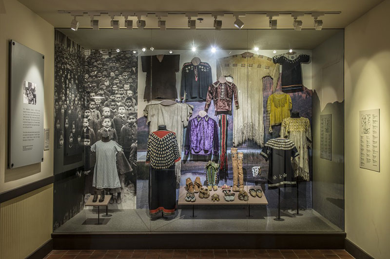 Display of children's clothing of various Native American tribes and early 20th C boarding school uniforms.