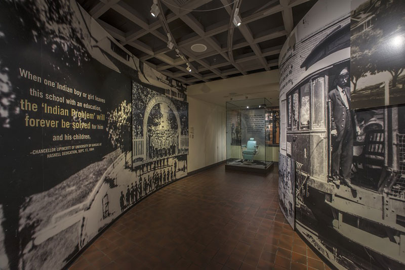 Curved walls with graphic imagery, text and photos of people, lead to a teal blue barber chair in a large glass enclosure