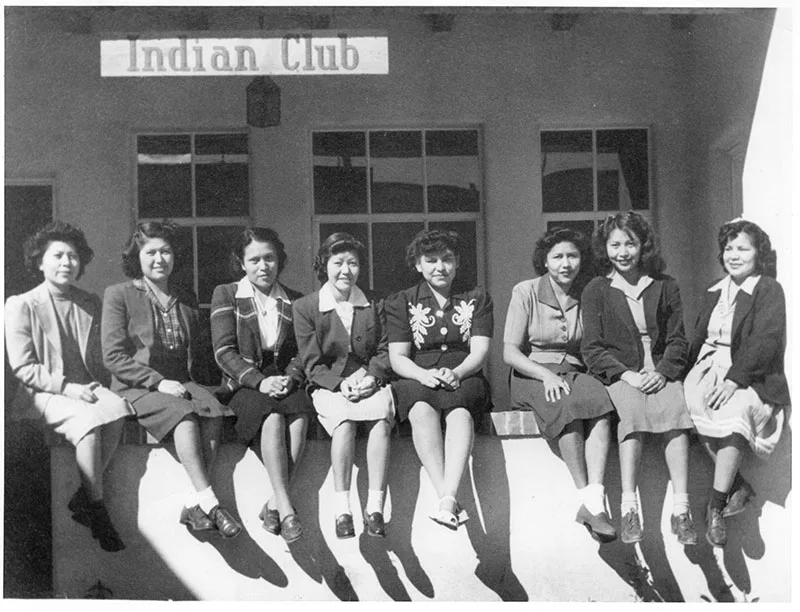 1930s black and white photo of young Native women in dresses and blazers sitting on a stucco wall posing for the camera in front of a veranda with a sign saying "Indian Club"