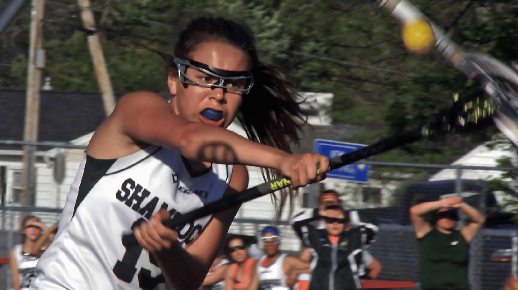 A lacrosse player in a white jersey and eye protection fiercely swings her stick during a game, with spectators watching in the background.