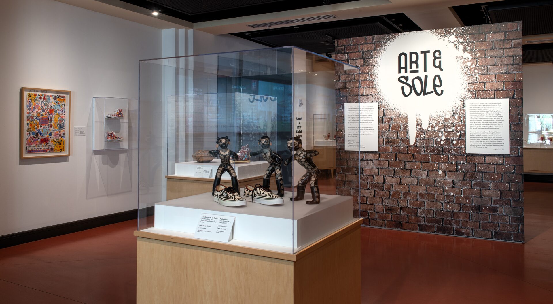 Museum exhibit titled "Art & Sole" featuring artwork and sculptures of sneakers displayed in glass cases and on walls, with descriptive panels and graffiti-style lettering on a brick wall.