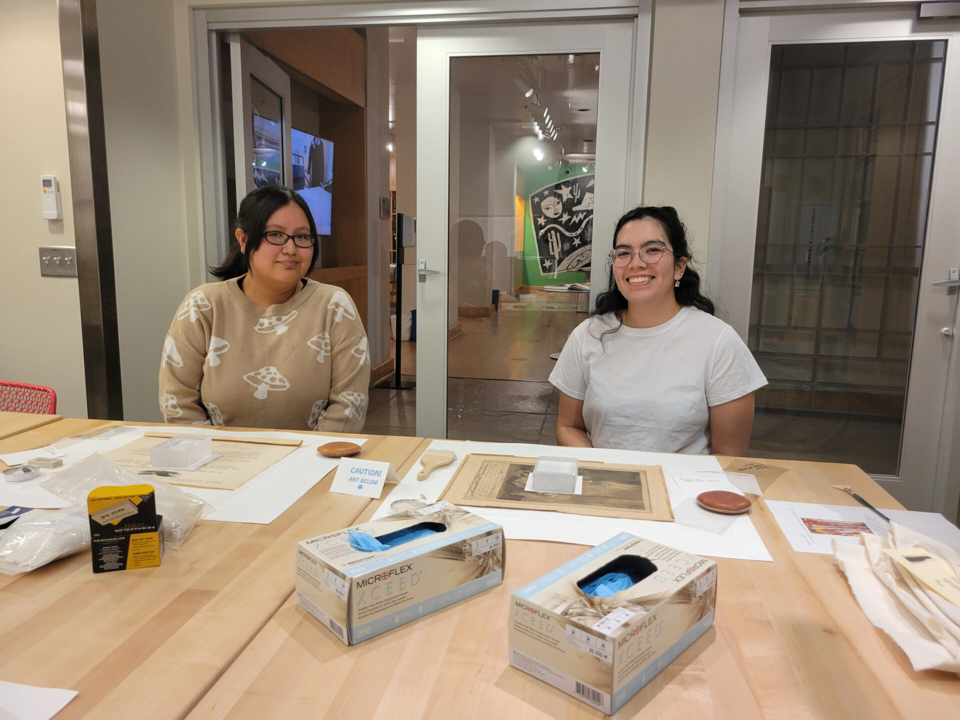 Two people sit at a table with various art supplies in a well-lit room. They are smiling and appear to be involved in an art-related activity. Boxes and materials are spread out on the table in front of them.