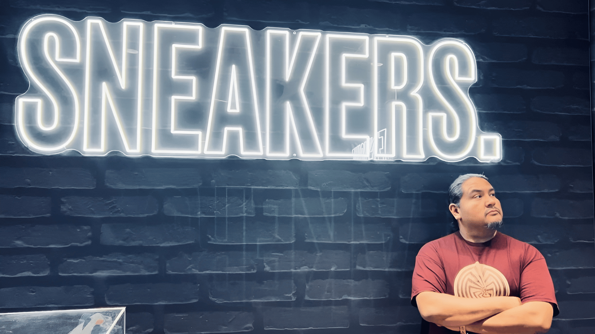 A person in a red t-shirt stands with arms crossed in front of a dark brick wall with a large, illuminated "SNEAKERS." sign.