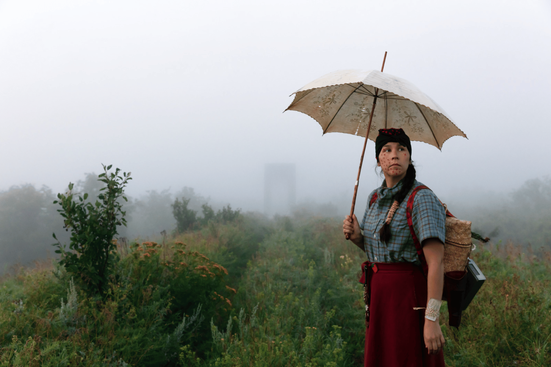 A person stands in a foggy field holding an ornate umbrella. They are wearing a plaid shirt, red skirt, and headscarf, with a basket strapped to their back and foliage in the background.