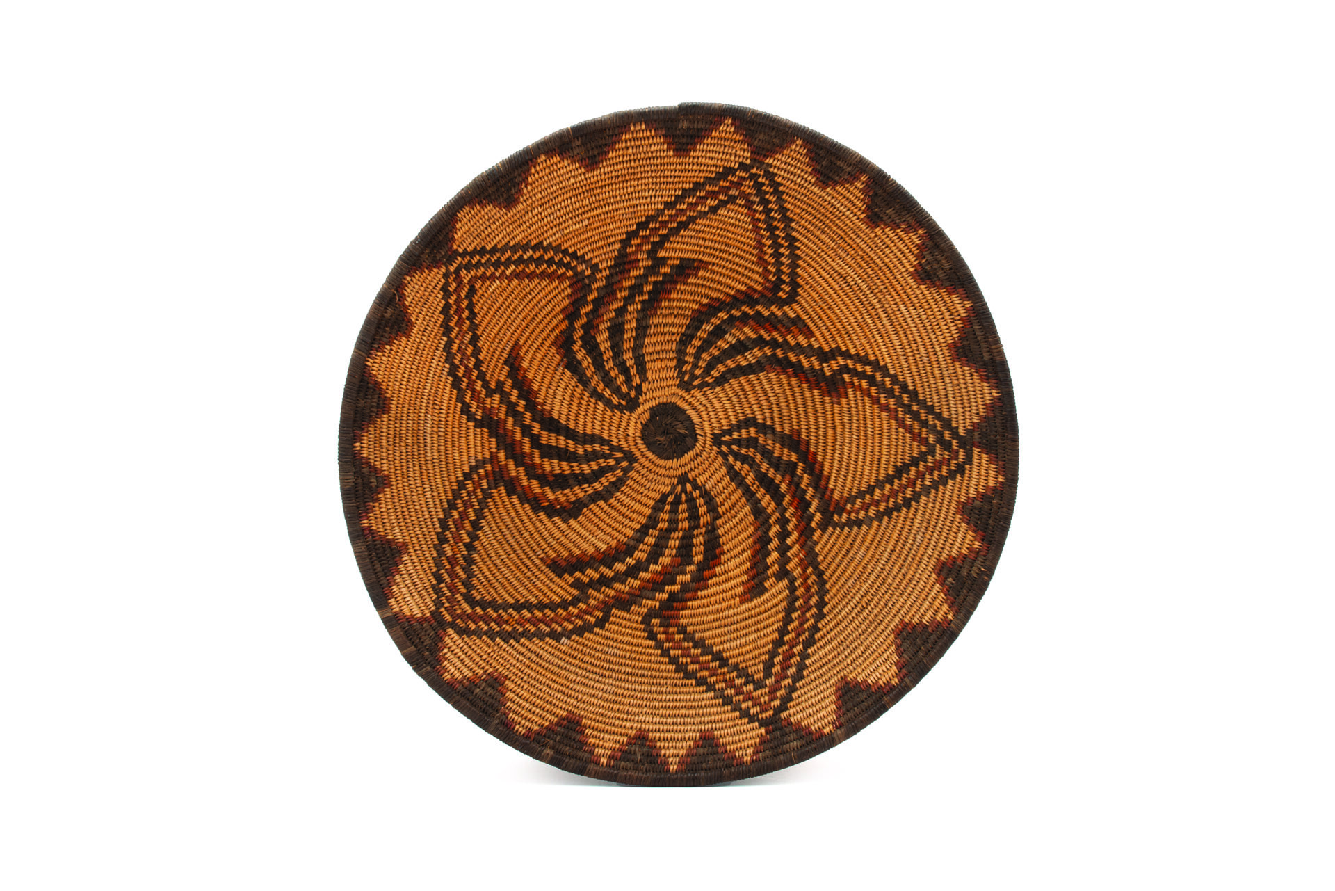 A round woven basket with intricate geometric patterns radiating from the center, featuring alternating triangular and swirling designs in various shades of brown.