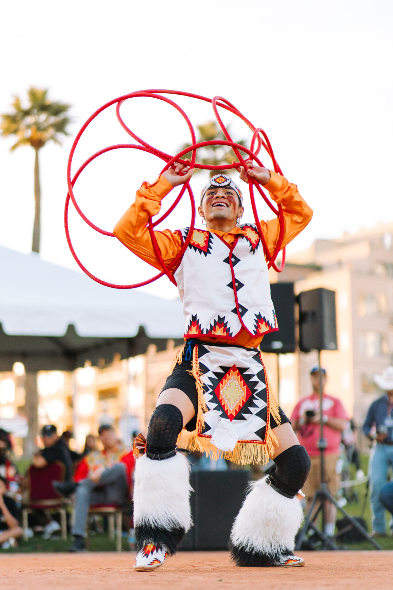 A person in traditional attire performs a hoop dance with multiple red hoops at an outdoor event. Other people are visible in the background.