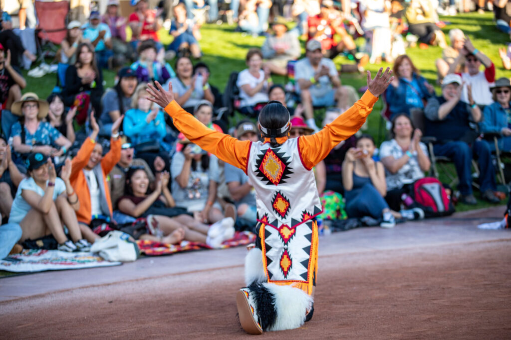 A person in traditional attire performs for a large, seated audience outdoors, with their arms raised and back to the camera. The audience watches attentively.