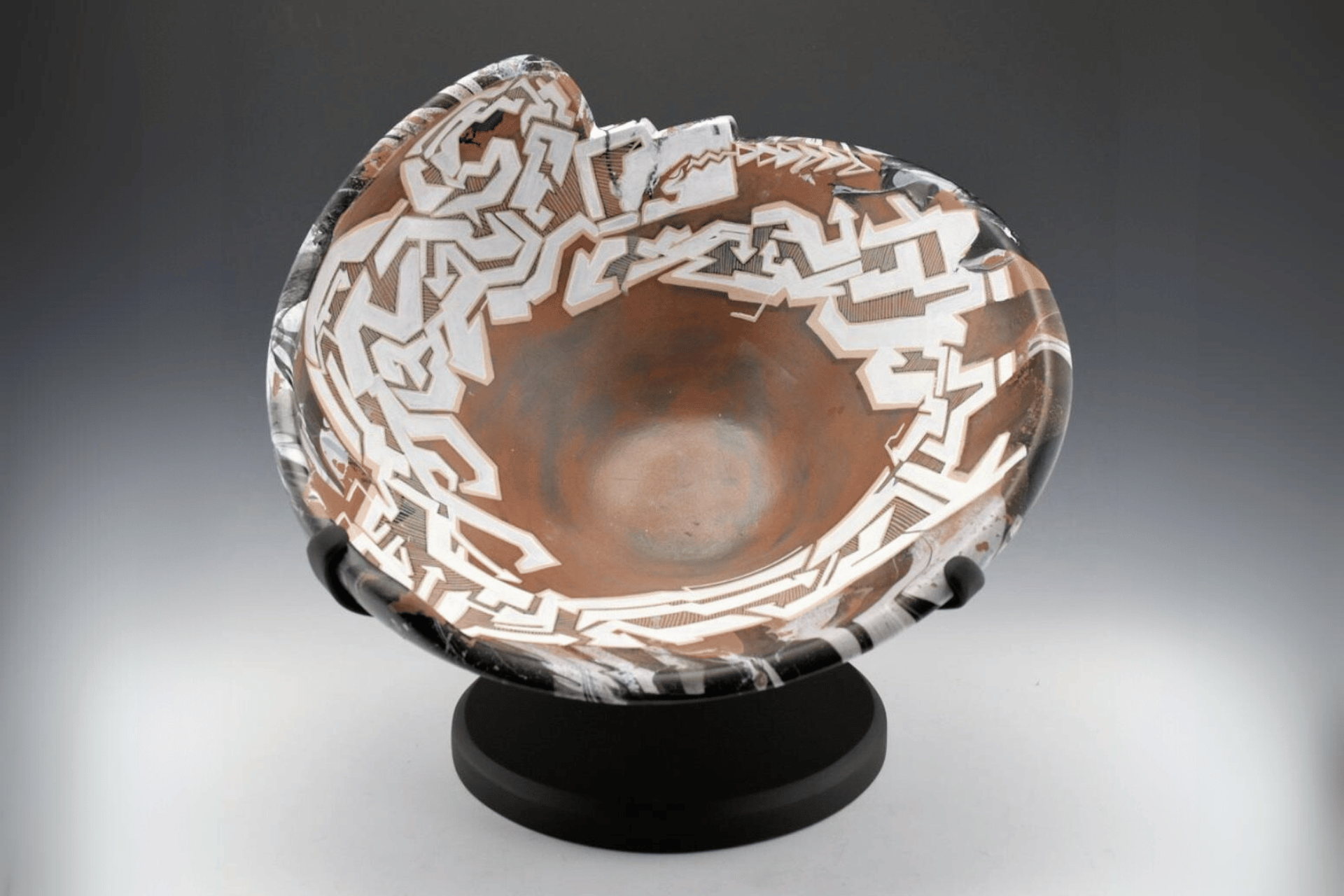 A ceramic bowl with intricate white geometric patterns on a brown and black background, displayed on a black stand.