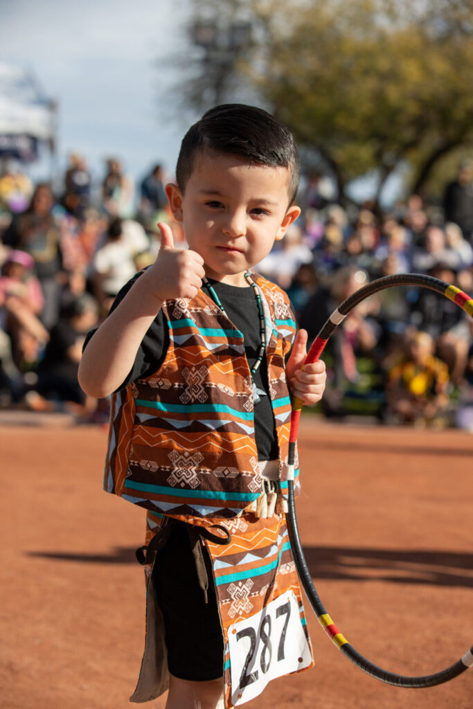 A young boy gives a thumbs-up while dressed in traditional attire, holding a hoop and wearing a number 287 tag, with a crowd in the background.