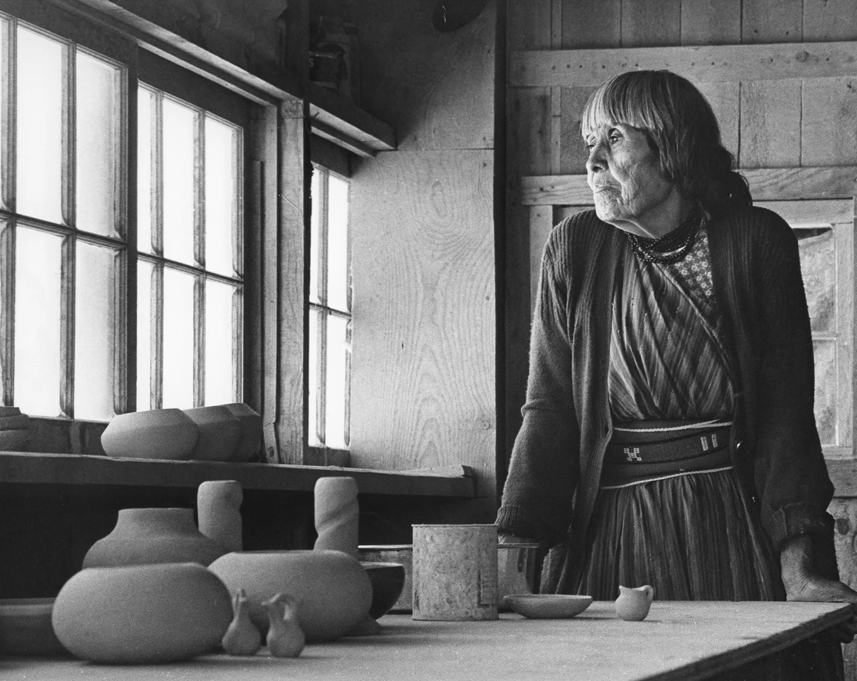An elderly person in a workshop gazes out the window, standing near a table with various pottery pieces.