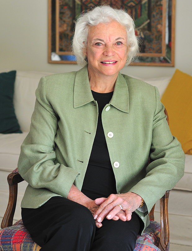 An elderly woman with white hair sits on a chair, wearing a green blazer over a black top. She's smiling with her hands resting on her lap, in a room with a couch and colorful cushions in the background.