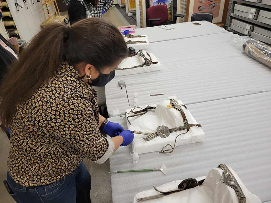 A woman wearing gloves and a mask examines historical artifacts laid out on a table in a storage room.