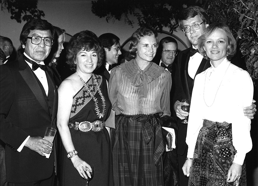 A group of men and women in formal attire pose for a photo at an event. One woman holds a drink, and another a clutch. People and greenery are visible in the background.