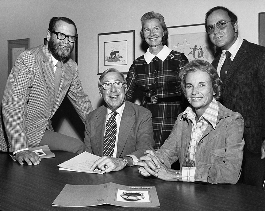 Five individuals gathered around a table, with two seated and three standing, all dressed in formal attire. Papers and a folder are on the table. Framed art hangs on the wall behind them.