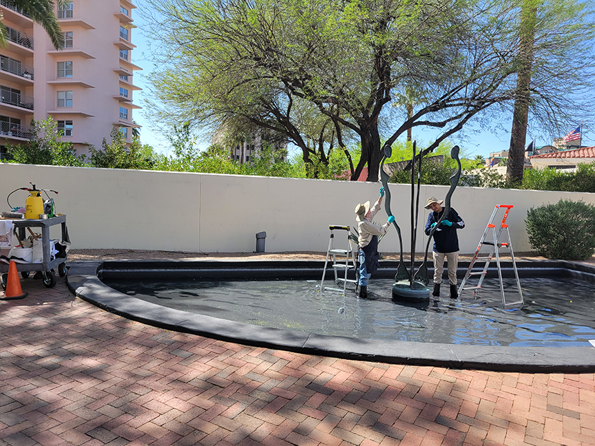 Two people work on a sculpture in a shallow water fountain, each standing on a step ladder. A maintenance cart is nearby, and buildings and trees are visible in the background.