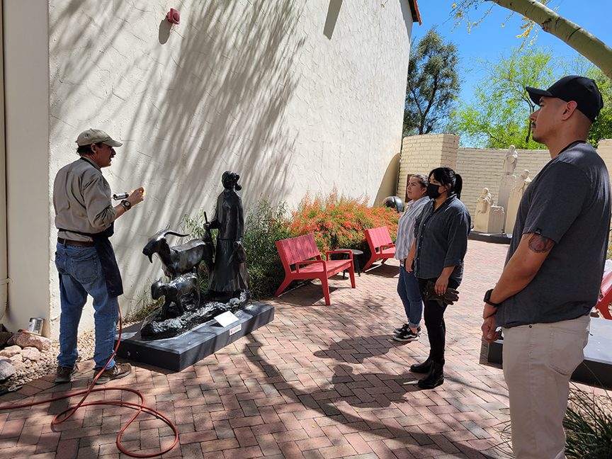 A group of people listens to a man giving a presentation about a black sculpture depicting various figures, outside near a white building with red benches.