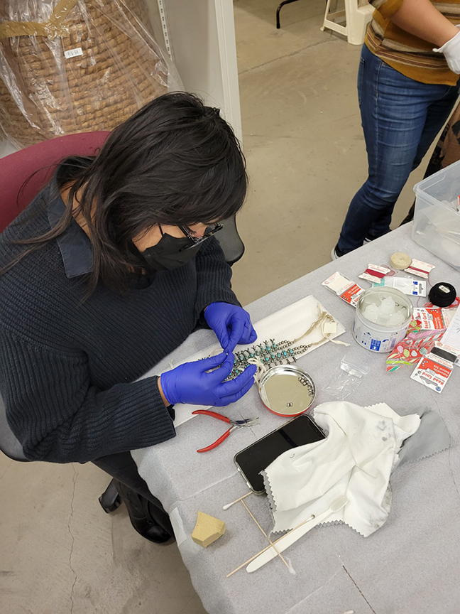 A person wearing a mask and gloves works on a delicate assembly at a table with various tools and materials, including a magnifying glass, phone, and small containers.