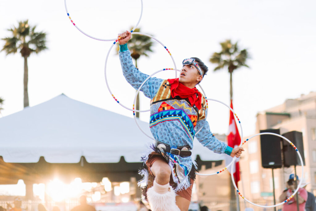 A man in traditional dress performs a hoop dance with multiple hoops, set against a backdrop of palm trees and a tent, during a cultural event at sunset.