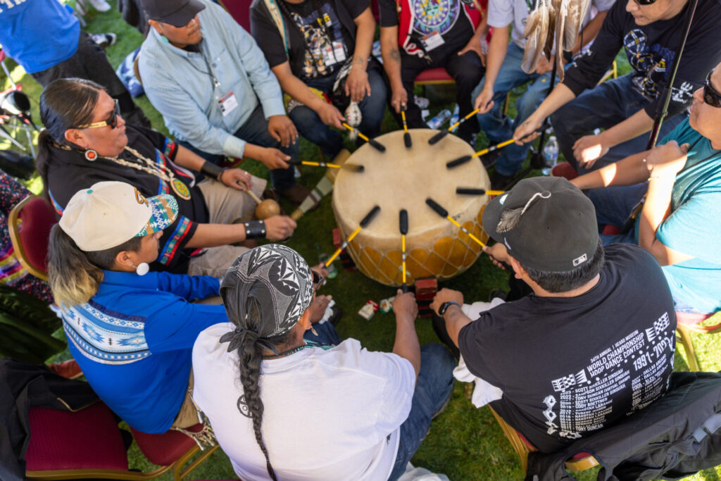 A group of people sit in a circle on chairs, playing a large drum with multiple drumsticks. They appear to be engaged in a cultural or musical activity outdoors.