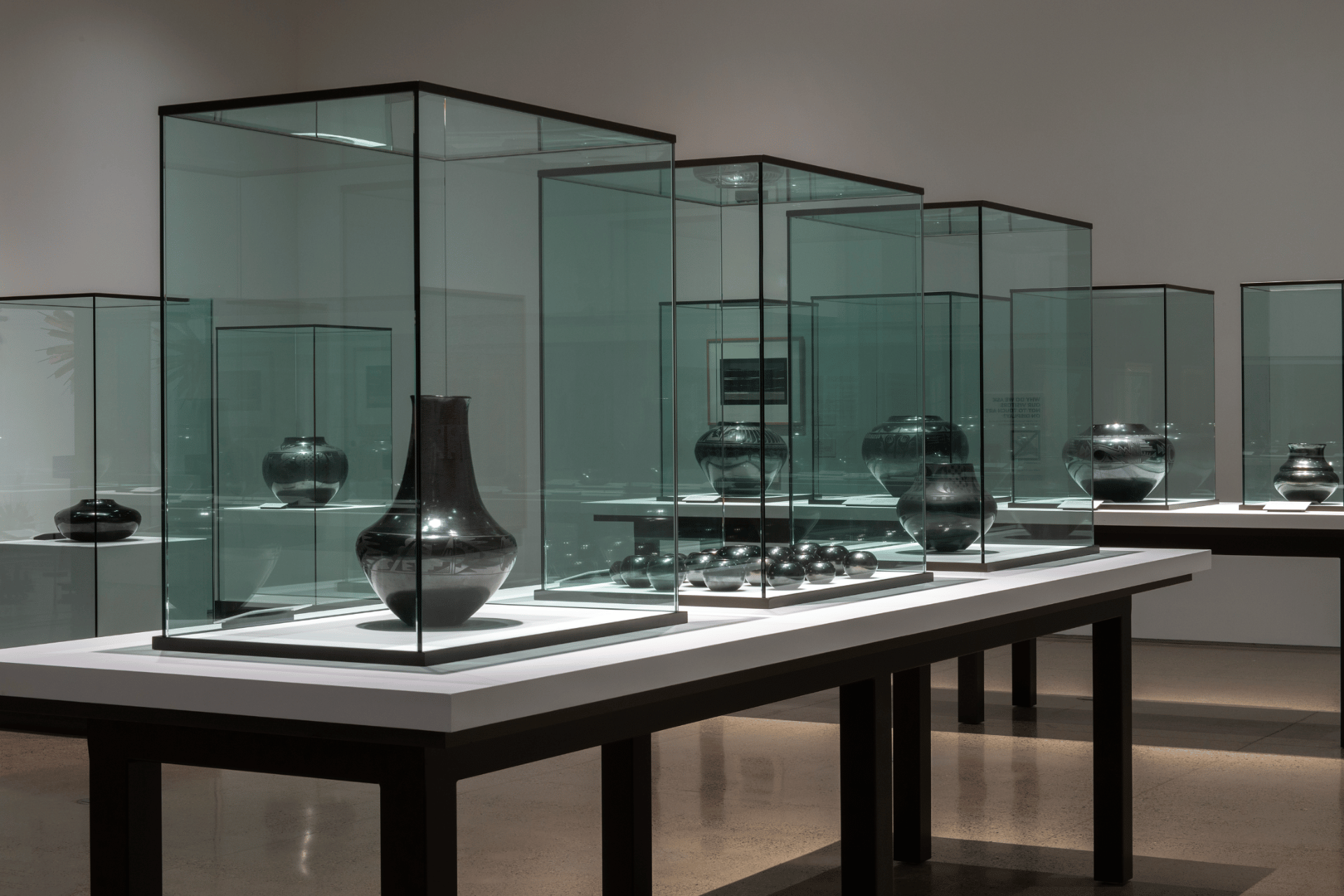 A museum exhibit displaying various black ceramic vases and bowls in glass cases, set on white platforms with a minimalist background.