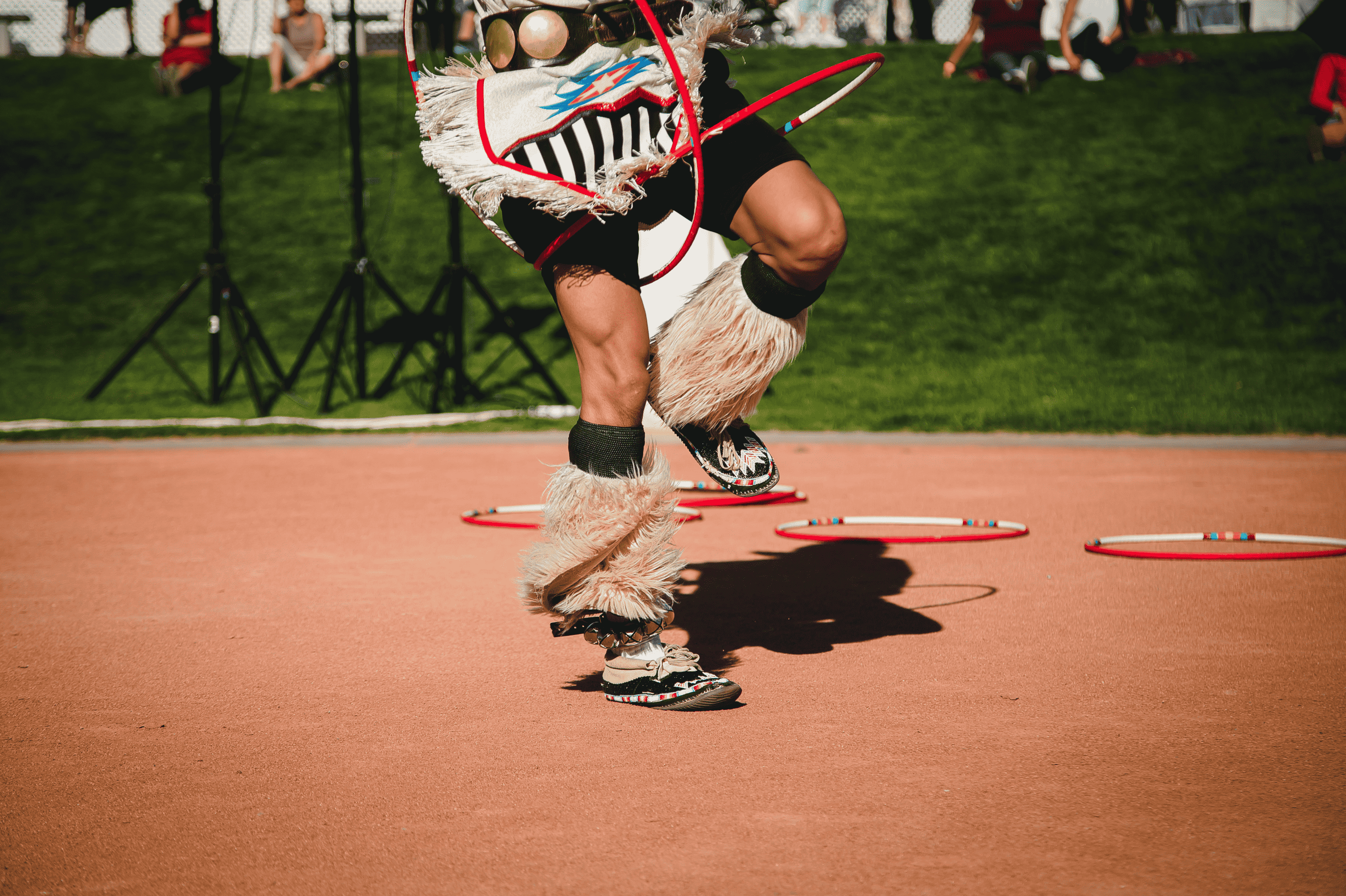 A person dances with hoops while wearing traditional attire, including fur-covered leggings and decorated shoes, on a sandy surface and grassy background.