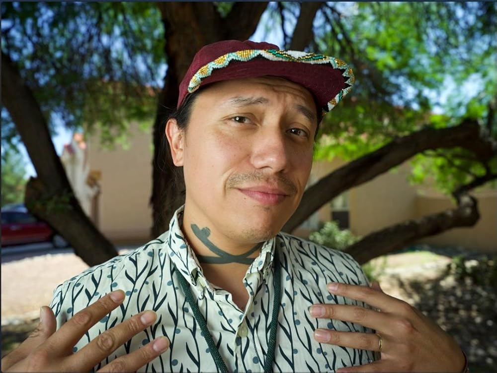 A person stands outdoors in front of a tree, wearing a maroon cap and a patterned shirt, with hands raised near their chest. There is a tattoo on their neck and a slight smile on their face.