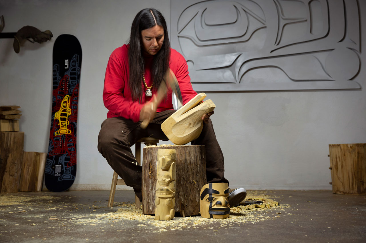 A person in a red shirt is carving a wooden sculpture while seated on a tree stump indoors. Nearby are completed carvings, wood shavings on the floor, and a decorated snowboard leaning against the wall.