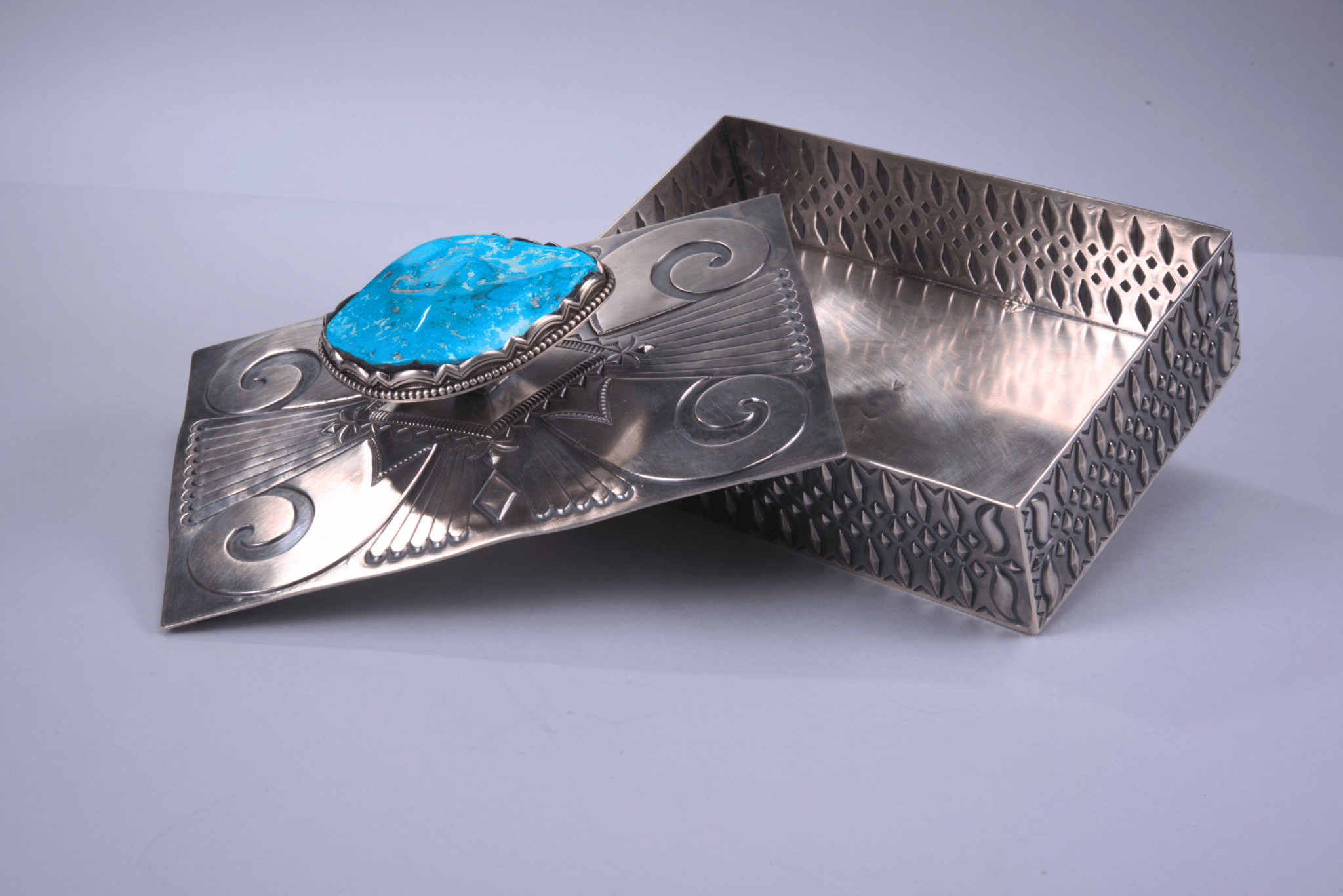 A silver decorative box with intricate patterns, partially opened to reveal its interior. The lid features an ornate turquoise stone setting.