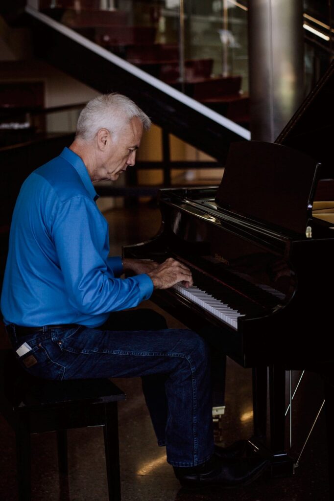 A man with gray hair in a blue shirt sits at a black grand piano, playing music.