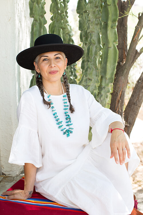 A woman in a white dress and black wide-brimmed hat sits near a cactus. She is wearing a turquoise necklace and has braids in her hair.