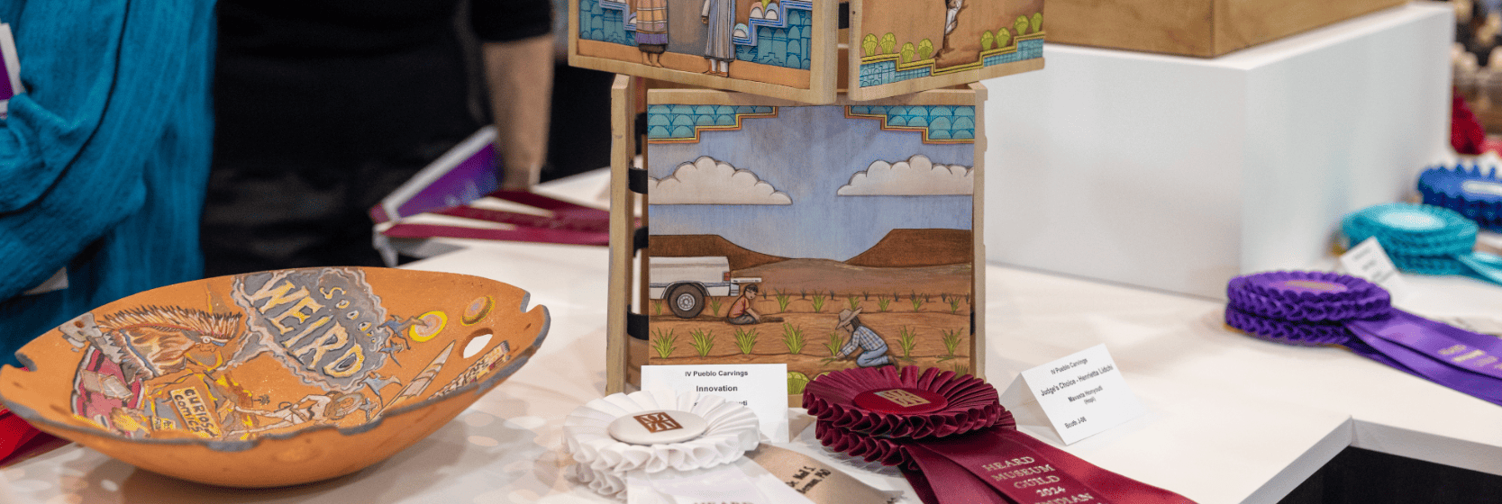 A display of various handcrafted art pieces with text labels, including a painted bowl and a tall box with scenic illustrations. Several ribbons are visible, indicating they won awards.