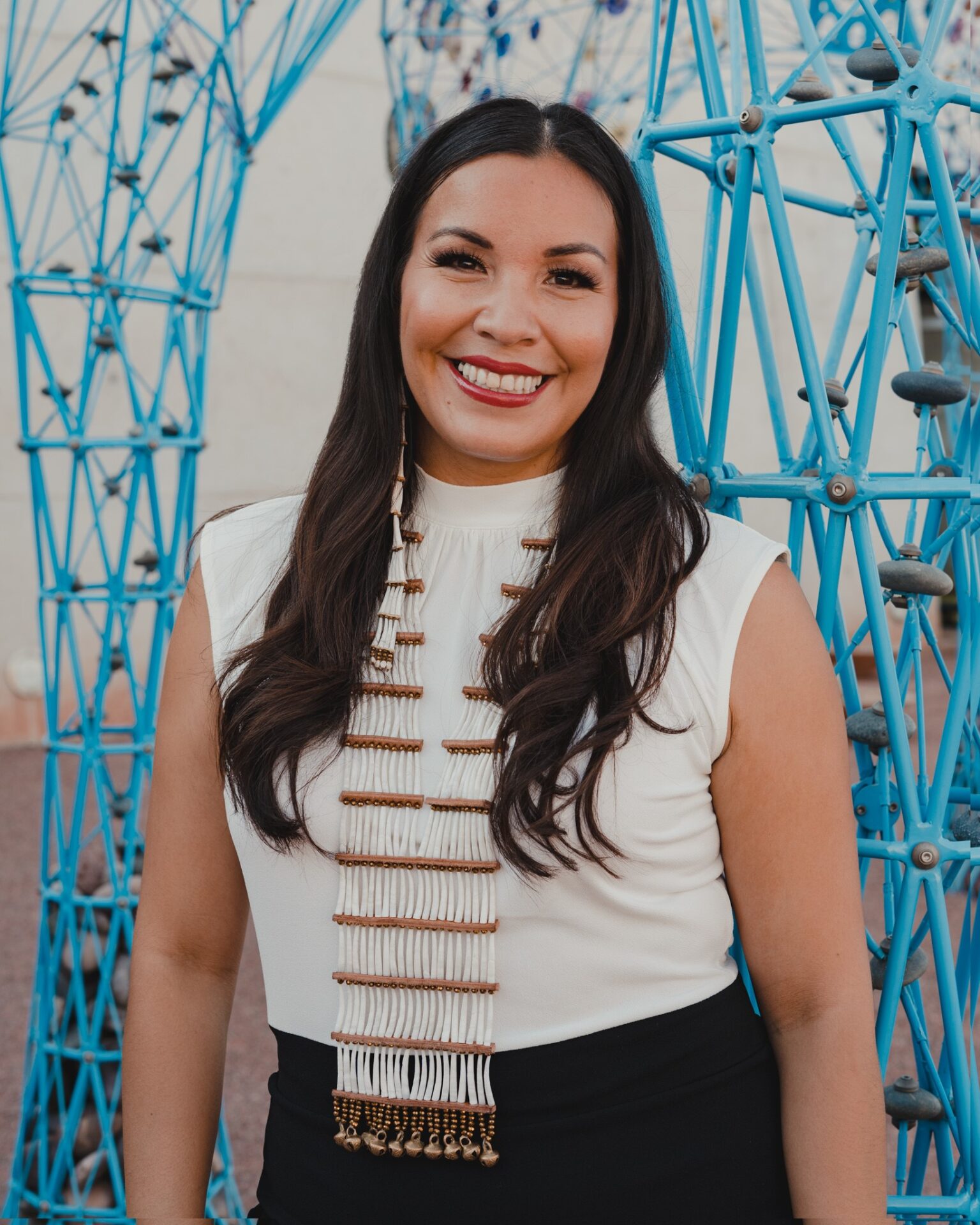 A woman with long dark hair wearing a white sleeveless top and a beaded necklace smiles in front of a blue geometric structure.