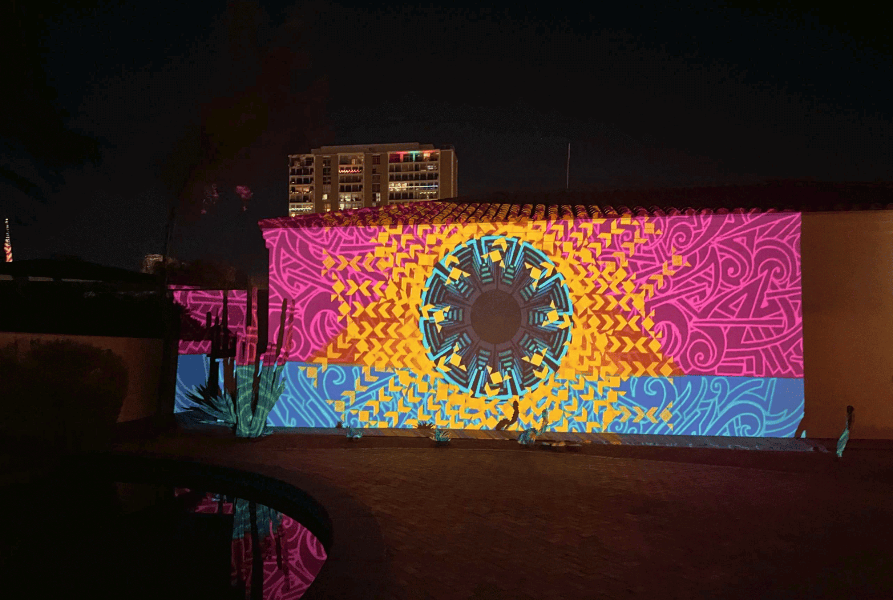 A building with an intricate, colorful projection of geometric and floral patterns on its facade is illuminated at night.