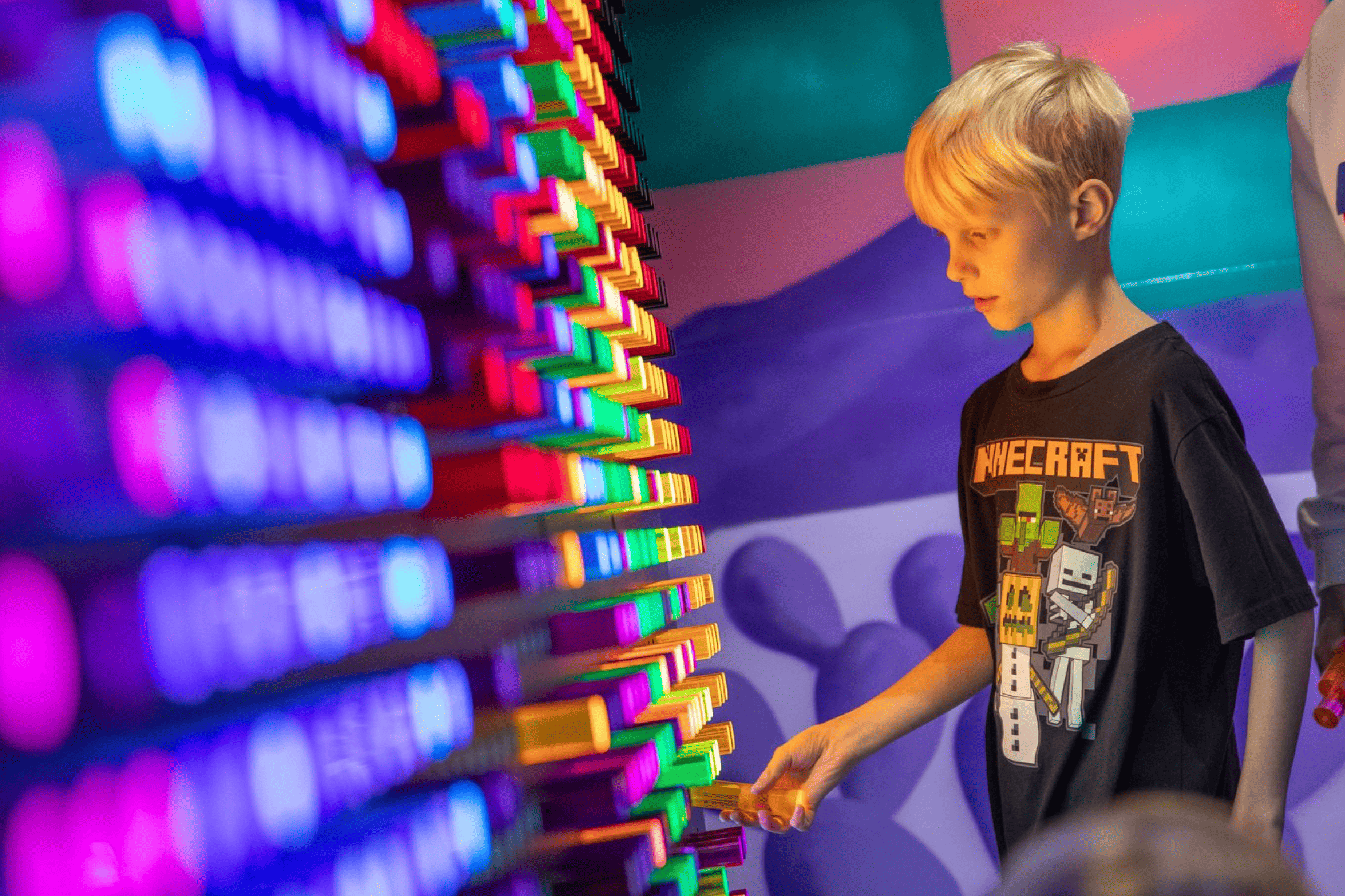 A young boy with blonde hair interacts with a light-up board featuring colorful pegs in a dark room. He is wearing a Minecraft-themed T-shirt and is focused on placing a peg.