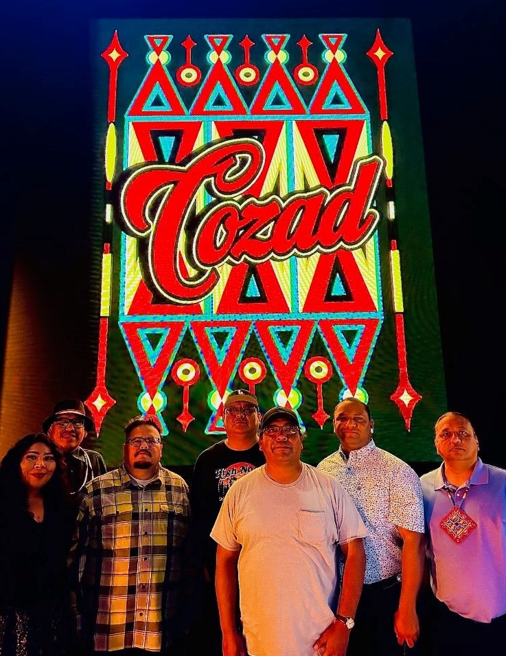 A group of seven people stands in front of an illuminated sign with vibrant patterns and the word "Cozad" in large letters.